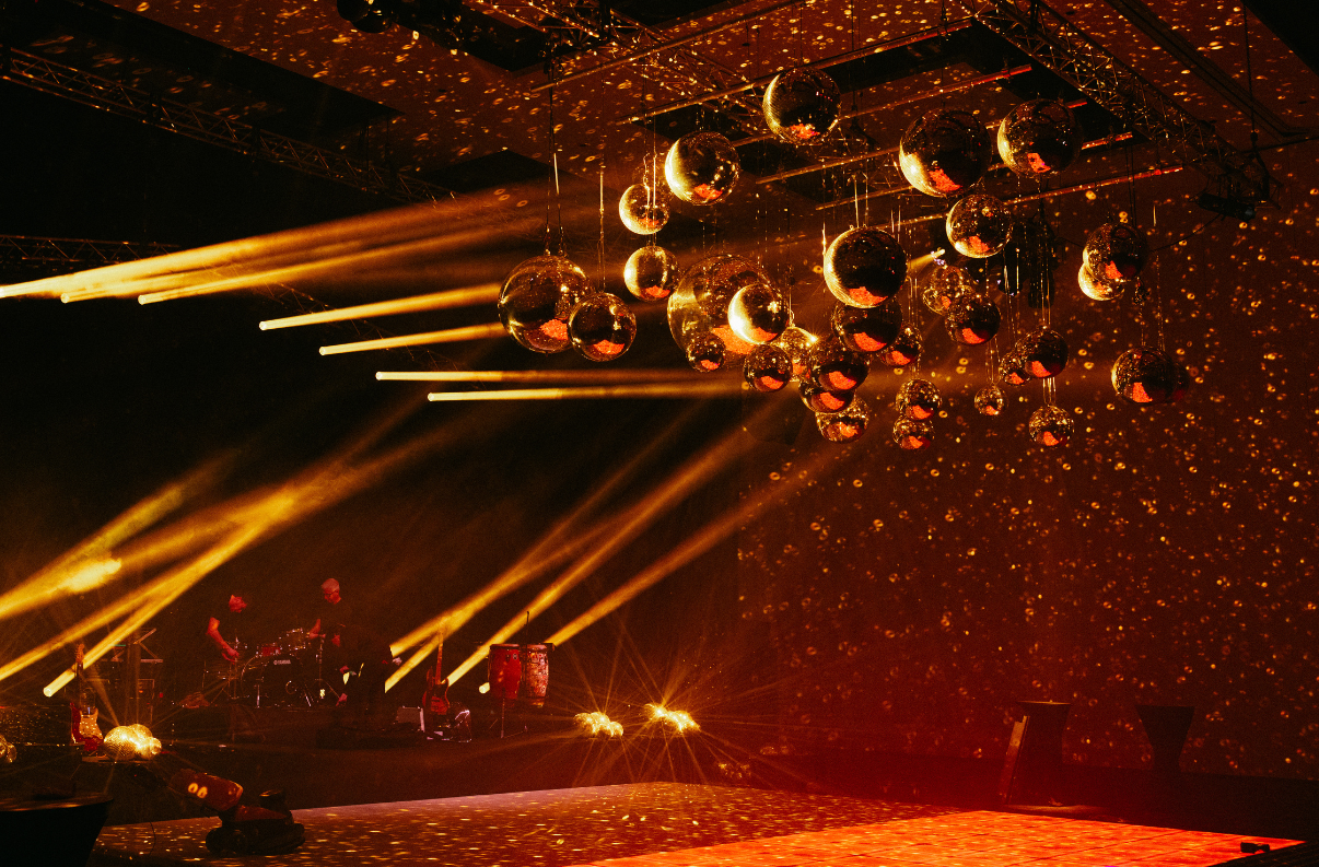 A stage-like performance space with multiple hanging disco-ball style mirrors, strong golden light beams, and a dark background with scattered reflective specks.